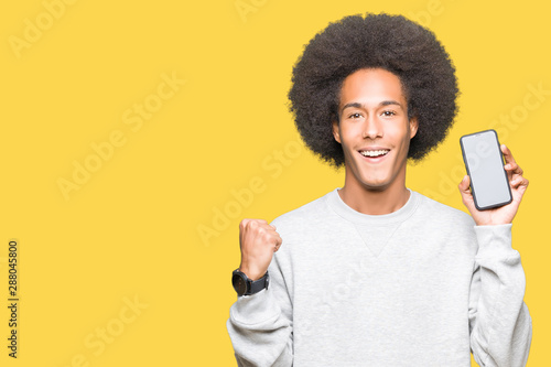 Young african american man with afro hair showing smartphone screen screaming proud and celebrating victory and success very excited, cheering emotion