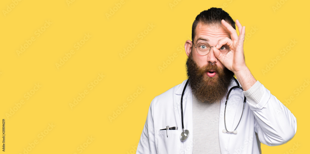 Young blond doctor man with beard wearing medical coat doing ok gesture shocked with surprised face, eye looking through fingers. Unbelieving expression. Young blond doctor man with beard wearing medical coat doing ok gesture shocked with surprised face, eye looking through fingers. Unbelieving expression.