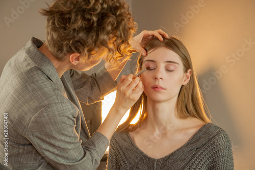 Makeup artist and hairdresser are preparing bride before wedding. Hairdresser-stylist with short stylish hairstyle applies makeup, makes false eyelashes, paints lips, curls girl, spectacular lighting