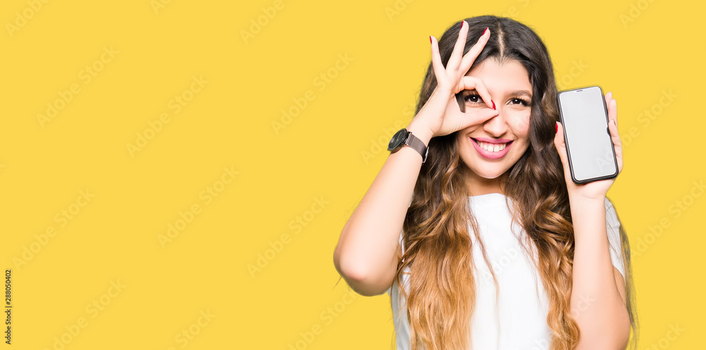 Young adult woman showing smartphone screen with happy face smiling doing ok sign with hand on eye looking through fingers - obrazy, fototapety, plakaty 