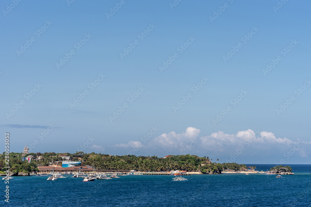Manoc-Manoc, Boracay, Philippines - March 4, 2019: Wide shot from sea of Cagban Jetty Port with tens of small boats. Dark blue see in front, green band of jungle trees with some red roofs under blue s