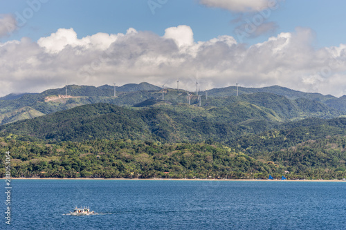 Caticlan, Malay, Philippines - March 4, 2019: Green mountain range with windmills dispersed under blue sky with thick gray cloudscape. Blue sea wth small outrigger boat. photo