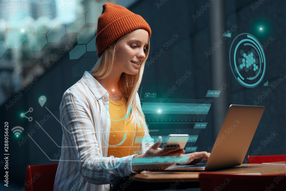Freelancers of the future could be working with a digital HUD around them. A millennial young woman is working with her laptop and phone in a cafe, while a holographic projection encircles her. - obrazy, fototapety, plakaty 