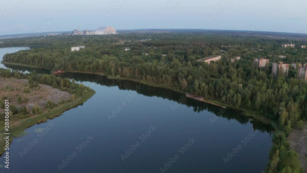 Aerial shot of quiet river and abandoned town with high-rise buildings among green trees, new safe confinement at nuclear power plant in distance, Pripyat, Chernobyl exclusion zone Aerial shot of quiet river and abandoned town with high-rise buildings among green trees, new safe confinement at nuclear power plant in distance, Pripyat, Chernobyl exclusion zone