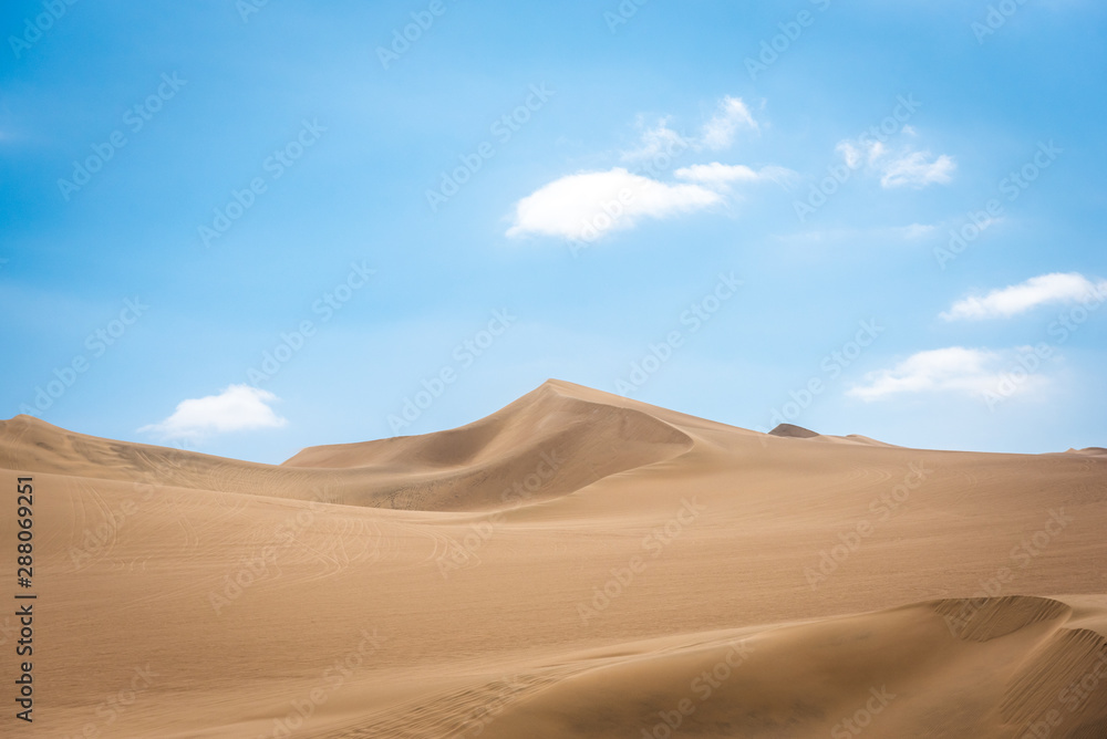 The swirling sand dunes of the Peruvian desert with light fluffy clouds above on a warm sunny day creating a spectacular contrast across the horizon