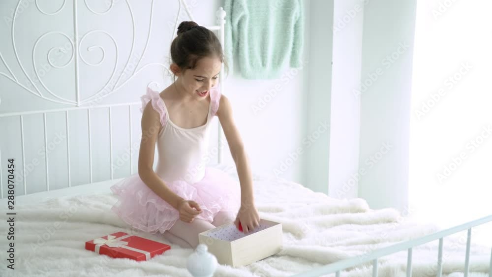4K Medium shot of happy beautiful little girl in pink ballet tutu and leotard sitting on the bed in bedroom and playing mini red heart. Cute child girl in pink ballet dress playing alone on the bed 4K Medium shot of happy beautiful little girl in pink ballet tutu and leotard sitting on the bed in bedroom and playing mini red heart. Cute child girl in pink ballet dress playing alone on the bed