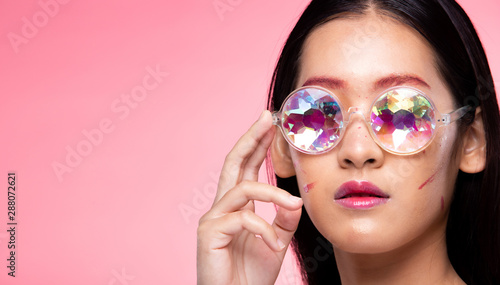 Korean Fashion Model looks at camera for shooting new collection Sunglasses. Beautiful Asian Woman with colorful trend make up wear Kaleidoscope Glasses with colorful, studio lighting pink background