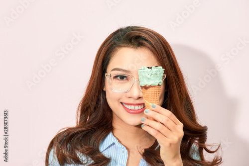 Young playful in eyeglasses covering eye with ice-cream cone and laughing excitedly on light background.