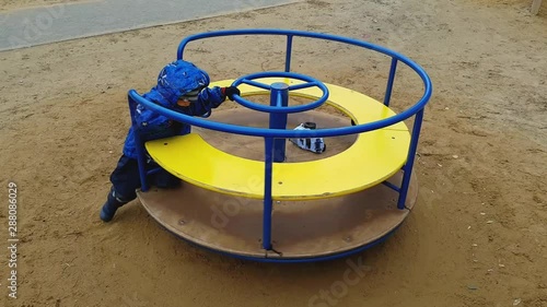 The little boy of 4 years in a jacket shakes himself on a merry-go-round in playground. Spring cloudy weather. Blue and yellow carousel. Top view.