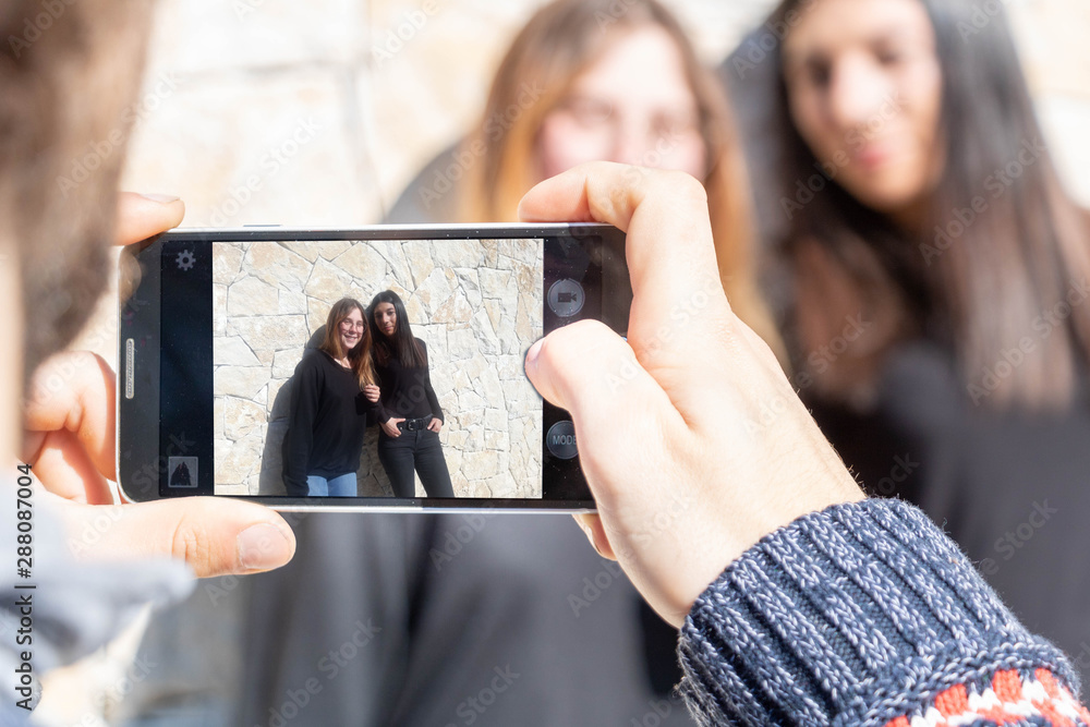 Deux jeunes femmes se faisant prendre en photo avec un smartphone par un ami - groupe multiethnique multiracial