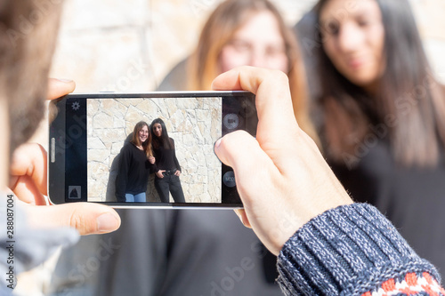 Deux jeunes femmes se faisant prendre en photo avec un smartphone par un ami - groupe multiethnique multiracial photo