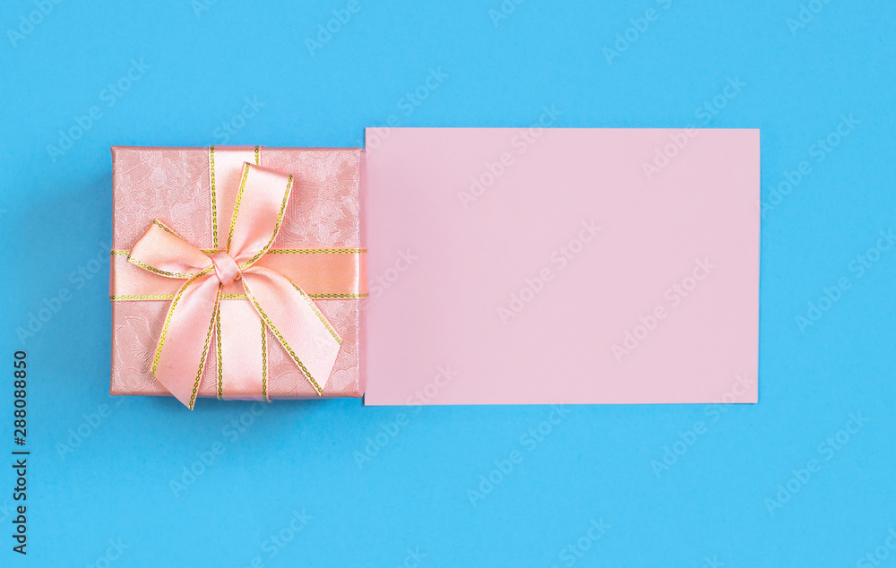 Pink gift box and blank card for writing on a blue background. Layout for recording. Flat lay, top view Pink gift box and blank card for writing on a blue background. Layout for recording. Flat lay, top view