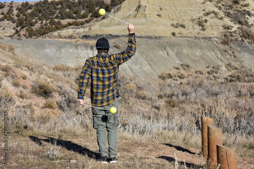 Hipster twirling two neon poi balls with back facing towards camera wearing a yellow and blue plaid long sleeve shirt, pants, and a backwards cap. Surrounded by rocky hills on a sunny but chilly day. photo