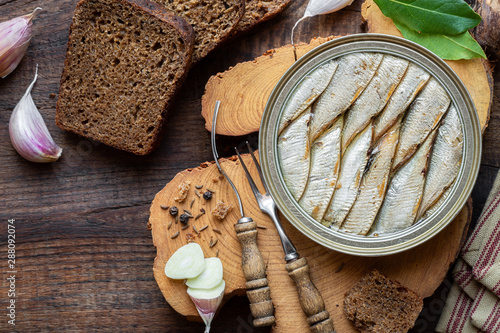 Obraz na płótnie Smoked sprat in a tin served with rye bread and garlic cloves on a rustic wooden cutting board