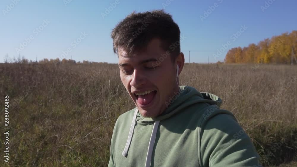 Smiling cute caucasian young man with thin stalk in mouth, wearing light-green hoody, is slowly walking alone in countryside along empty field on clear autumn day with blue sky. Smiling cute caucasian young man with thin stalk in mouth, wearing light-green hoody, is slowly walking alone in countryside along empty field on clear autumn day with blue sky.