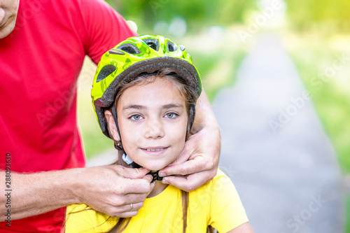 Father puts a helmet on his daughter in summer park. Empty space for text © Ermolaev Alexandr