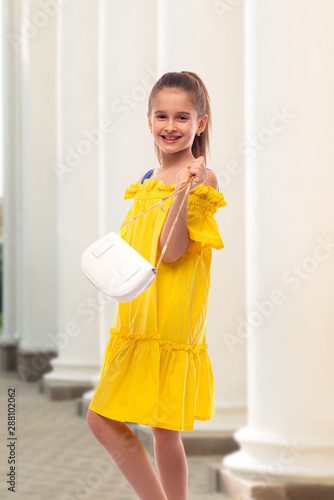 Portrait of a positive girl with long hair  and holding a small white handbag in her hand.  She walks past classic white columns