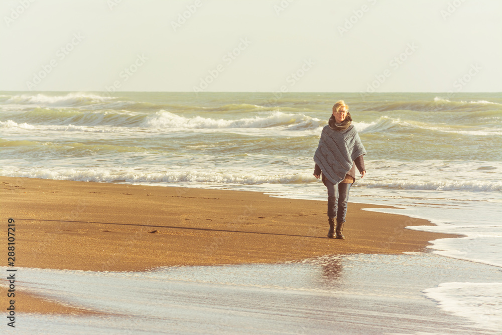Mature woman with poncho that walks on the seashore in autumn - obrazy, fototapety, plakaty 