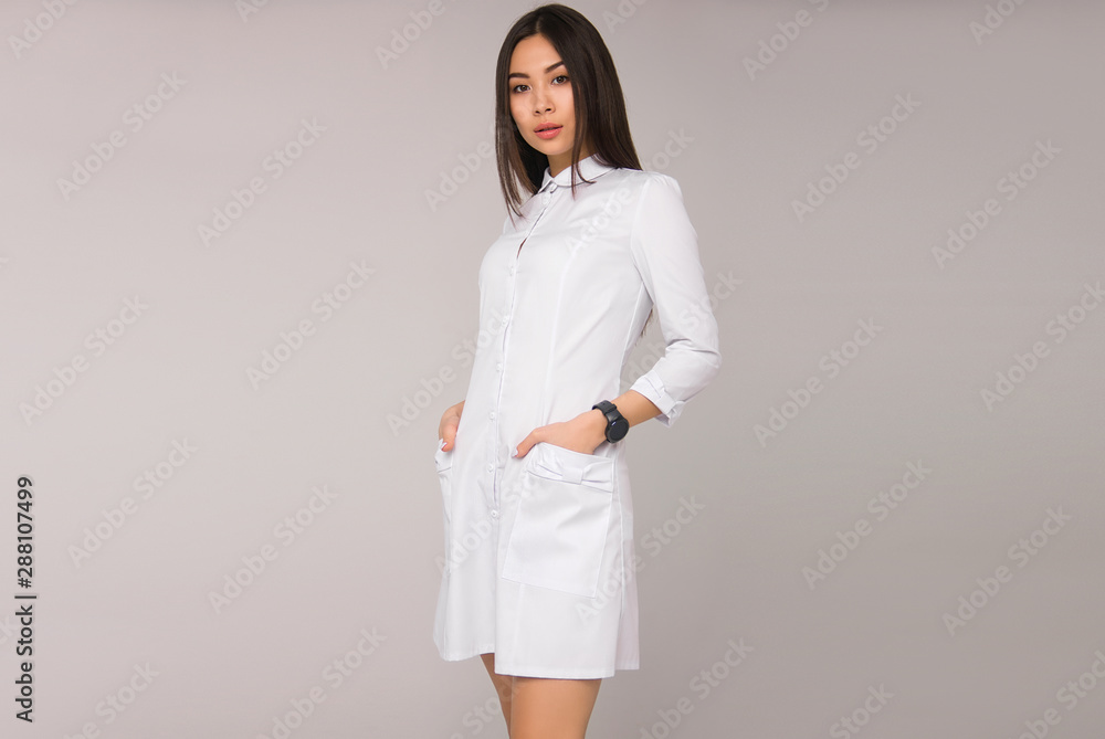 young stylish nurse with long brunette hair in white medical dress is standing with hands in her pockets, looking straight and smiling on the white wall background. medical fashion concept, free space young stylish nurse with long brunette hair in white medical dress is standing with hands in her pockets, looking straight and smiling on the white wall background. medical fashion concept, free space