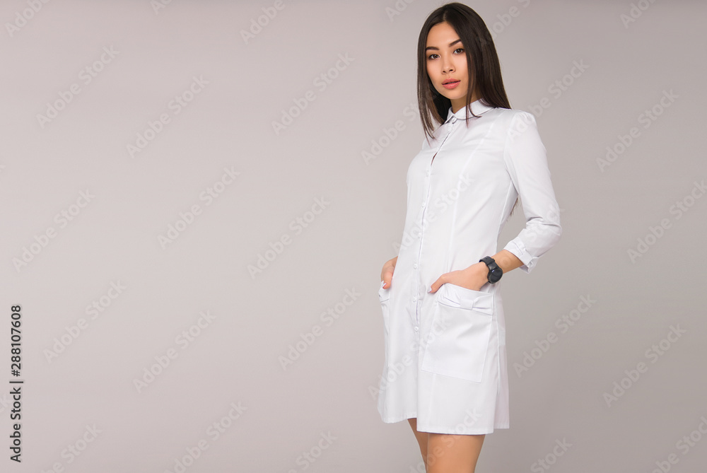 young stylish nurse with long brunette hair in white medical dress is standing with hands in pockets, looking straight and smiling on a white wall background. medical concept, free space on left side young stylish nurse with long brunette hair in white medical dress is standing with hands in pockets, looking straight and smiling on a white wall background. medical concept, free space on left side