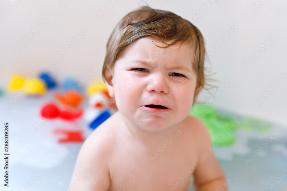 Cute adorable sad baby girl taking foamy bath in bathtub. Crying toddler playing with bath rubber toys Cute adorable sad baby girl taking foamy bath in bathtub. Crying toddler playing with bath rubber toys