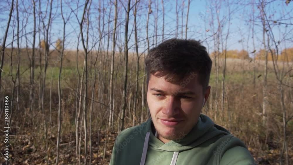 Good-looking caucasian young man, wearing light-green hoody is slowly walking alone in country side on narrow road covered with green high grass along empty wheat fields on clear day with blue sky. Good-looking caucasian young man, wearing light-green hoody is slowly walking alone in country side on narrow road covered with green high grass along empty wheat fields on clear day with blue sky.