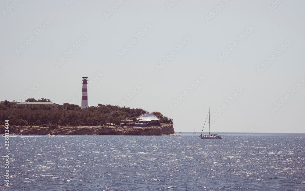 View of the coastal lighthouse on a cloudless day. The yacht drifting near a cliff on a boat trip.