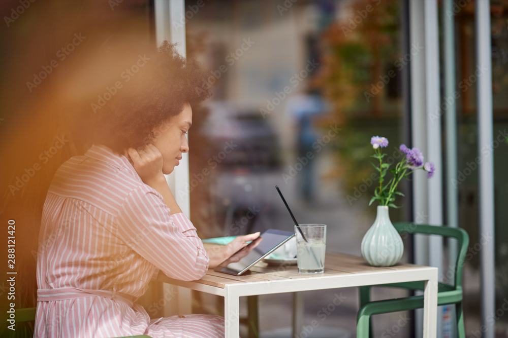 Grogeous mixed race woman in pink striped dress sitting in cafe and using tablet. On table are cup of coffee, book and lemonade. Grogeous mixed race woman in pink striped dress sitting in cafe and using tablet. On table are cup of coffee, book and lemonade.