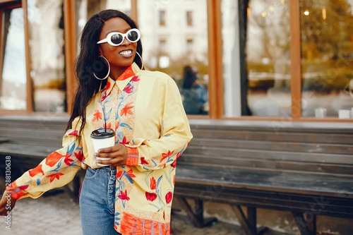 A beautiful and stylish black girl with long dark hair dressed in a yellow shirt and with sunglasses standing a summer city with coffee