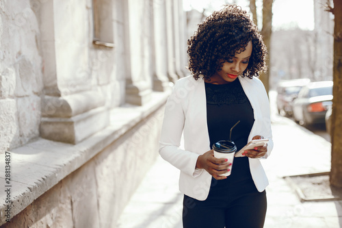 elegant and stylish dark-skinned girl with curly hair and in a white jacket walking around the summer city with coffee and using the phone