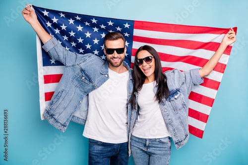 Portrait of cheerful married people in eyewear eyeglasses holding us flag screaming wearing denim jeans jacket isolated over blue background