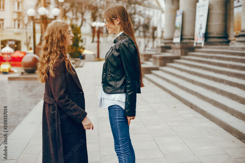 a beautiful stylish young girl with long curly hair and a long coat walking in the autumn city with her girlfriend in a black leather jacket and glasses