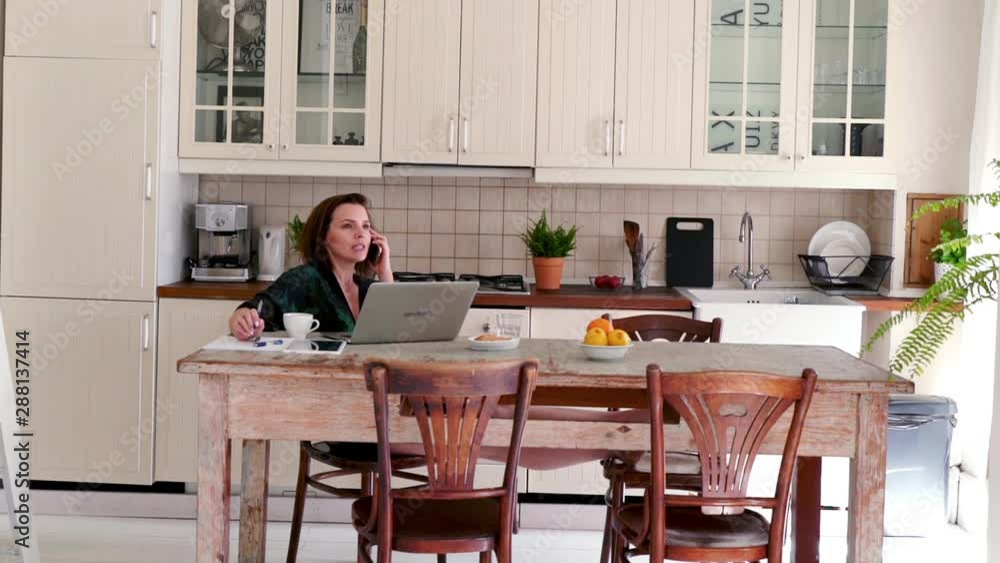 Young woman in bathrobe talking on cellphone and doing notes by table in kitchen Young woman in bathrobe talking on cellphone and doing notes by table in kitchen