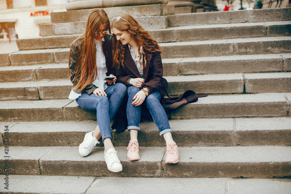 a beautiful stylish young girl with long curly hair and a long coat sitting in the autumn city with her girlfriend in a black leather jacket and glasses and they uses the phone a beautiful stylish young girl with long curly hair and a long coat sitting in the autumn city with her girlfriend in a black leather jacket and glasses and they uses the phone