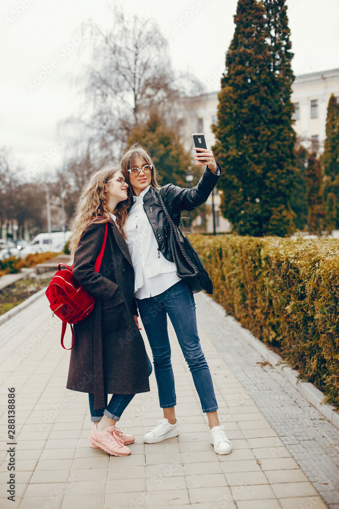 a beautiful stylish young girl with long curly hair and a long coat standing in the autumn city with her girlfriend in a black leather jacket and glasses and they uses the phone