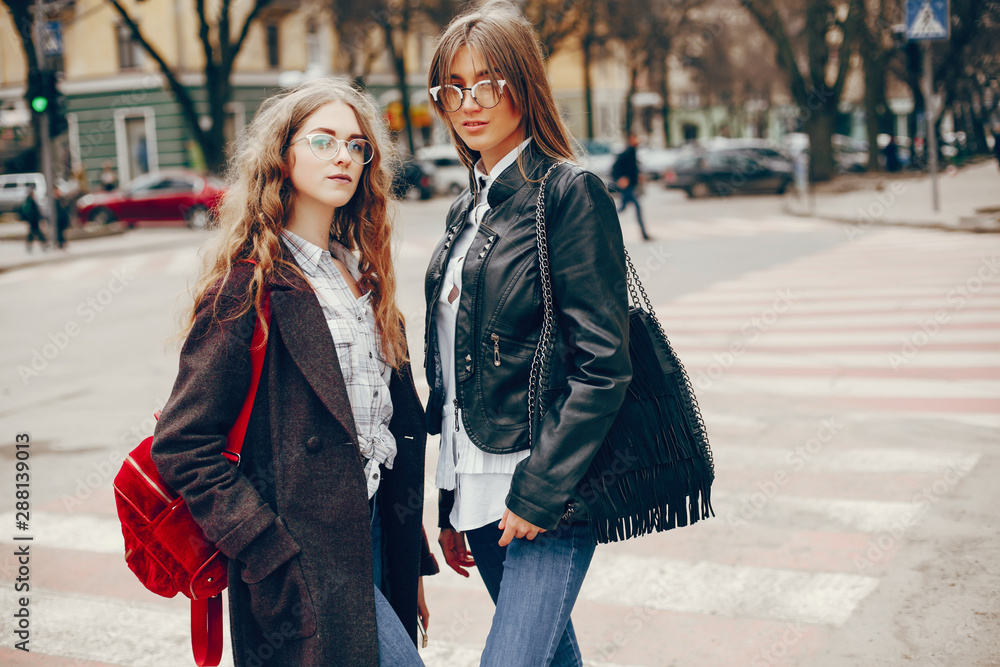 a beautiful stylish young girl with long curly hair and a long coat walking in the autumn park with her girlfriend in a black leather jacket and glasses