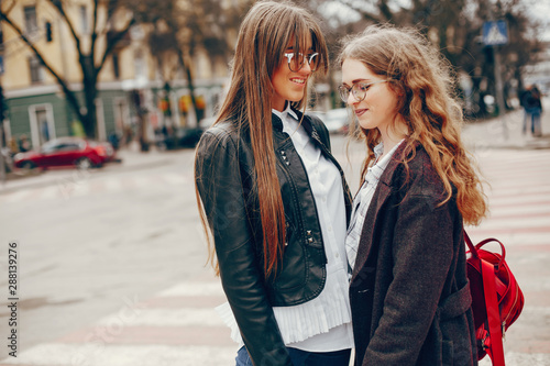 a beautiful stylish young girl with long curly hair and a long coat walking in the autumn park with her girlfriend in a black leather jacket and glasses