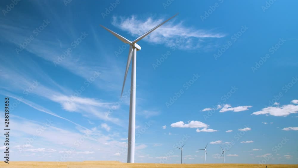 Hero shot of a wind turbine spinning against a blue sky with clouds over a yellow wheat field with three more turbines far in the distance Hero shot of a wind turbine spinning against a blue sky with clouds over a yellow wheat field with three more turbines far in the distance