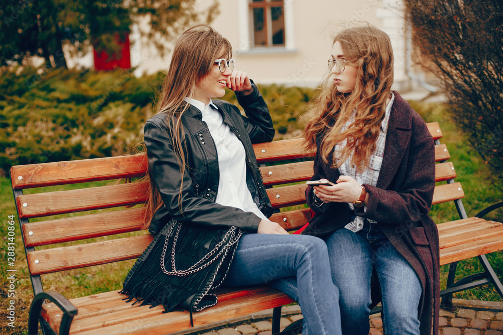 a beautiful stylish young girl with long curly hair and a long coat sitting in the autumn city with her girlfriend in a black leather jacket and glasses