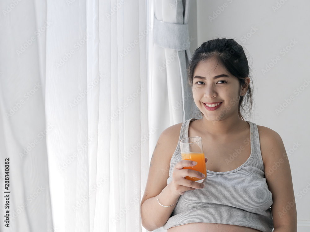Asian pregnant woman holding a glass of orange juice and standing near window, lifestyle concept. Asian pregnant woman holding a glass of orange juice and standing near window, lifestyle concept.