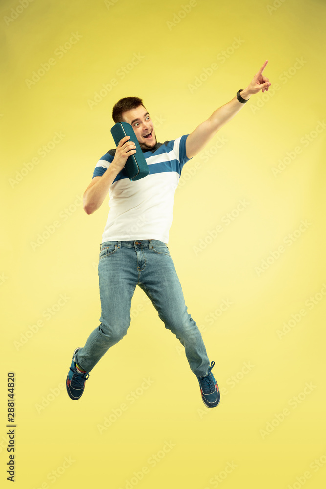 Sky sound. Full length portrait of happy jumping man with gadgets on yellow background. Modern tech, freedom of choices concept, emotions concept. Using portable speaker like superhero in flight.