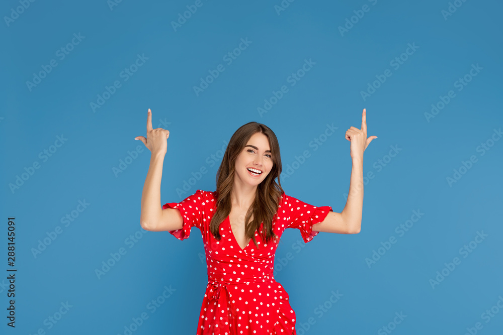 Attractive young woman with bright smile in red dress pointing fingers up over blue background. Attractive young woman with bright smile in red dress pointing fingers up over blue background.