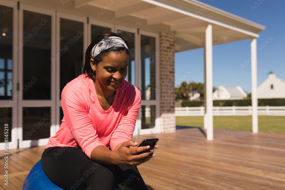 Fototapeta premium Woman using mobile phone while sitting on exercise ball in the backyard
