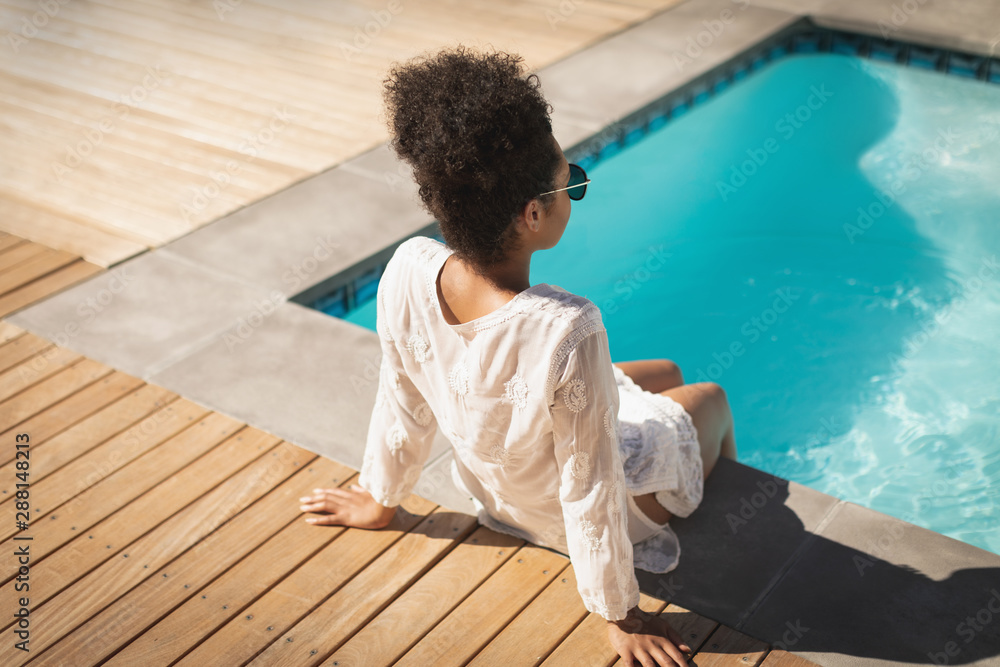 Young mixed-race woman with sunglasses sitting at poolside 