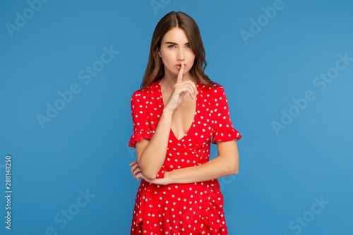 Charming young woman in red dress show keep silent gesture with serious face over blue background. © Screaghin