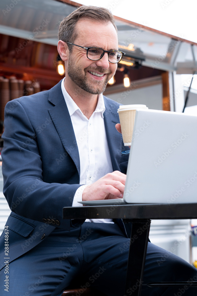 Mature Businessman Working On Laptop By Outdoor Coffee Shop Drinking Takeaway Coffee Mature Businessman Working On Laptop By Outdoor Coffee Shop Drinking Takeaway Coffee