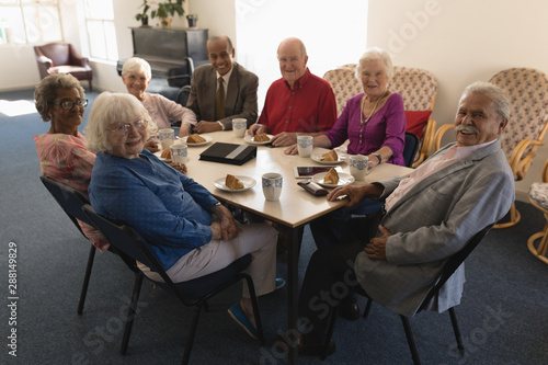 Front view of happy group of senior friends sitting on dining table and looking at camera