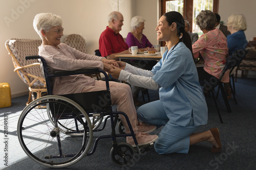 Side view of female doctor holding hands and talking with disable senior woman at home