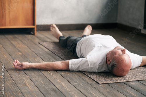 Senior caucasian man meditating on a wooden floor of living room and lying in Shavasana pose after practice. Yoga relax, meditating concept, close up