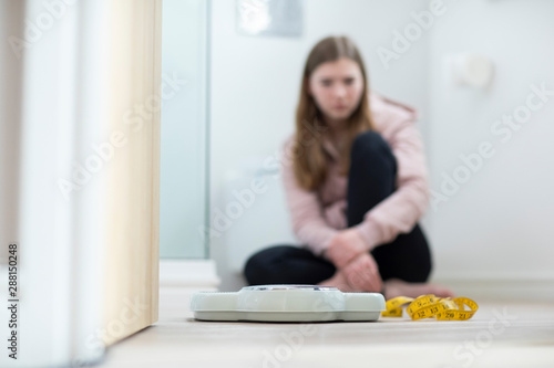 Unhappy Teenage Girl Sitting In Bathroom Looking At Scales And Tape Measure photo